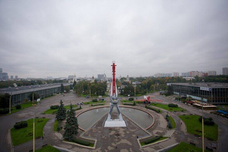 Вид сверху на площадь с круглым бассейном и макетом ракеты в центре; по сторонам стоят выставочные здания.