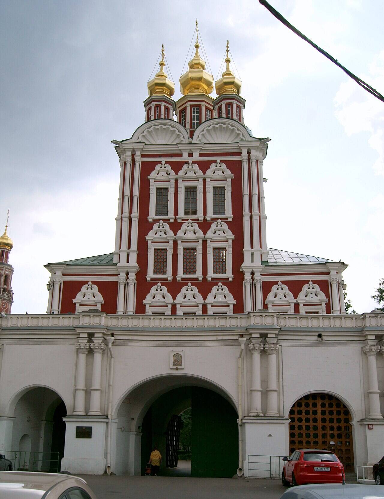 Красно-белая церковь с золочёными главами над тремя арками монастырских ворот; внизу стоит автомобиль.
