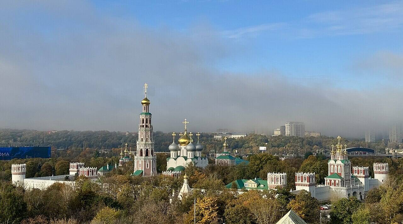 Вид сверху на Новодевичий монастырь среди деревьев: высокая колокольня, пятиглавый собор, стены и башни.