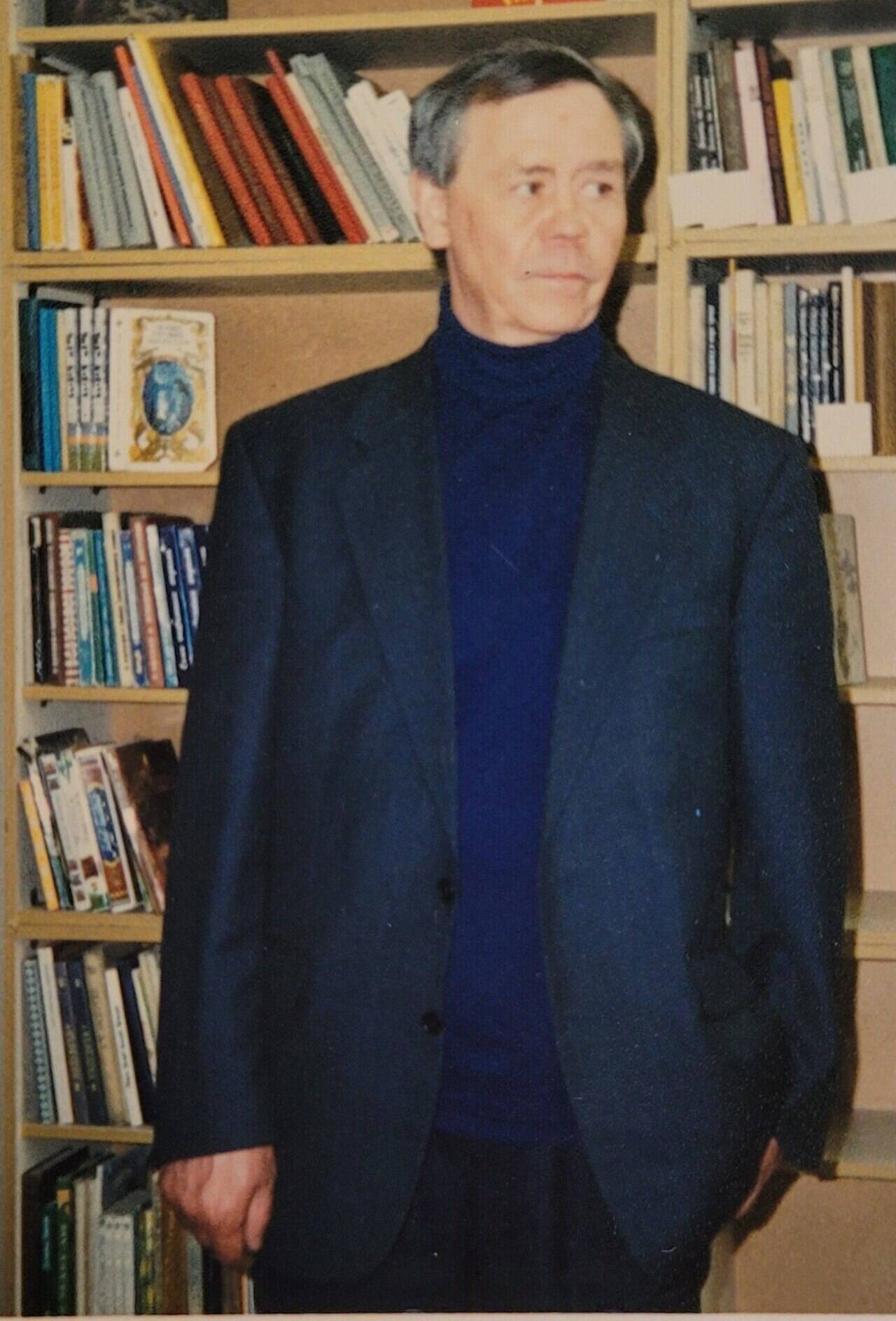 Валентин Распутин в тёмном пиджаке стоит перед книжными стеллажами
