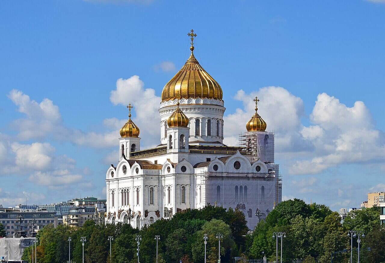 The white Cathedral of Christ the Saviour with its golden domes behind the trees; scaffolding is visible on the building to the right.