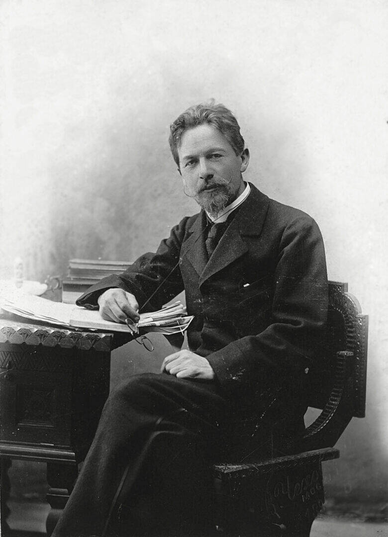 Anton Chekhov, wearing a dark suit, sits at a table with papers and looks at the camera.
