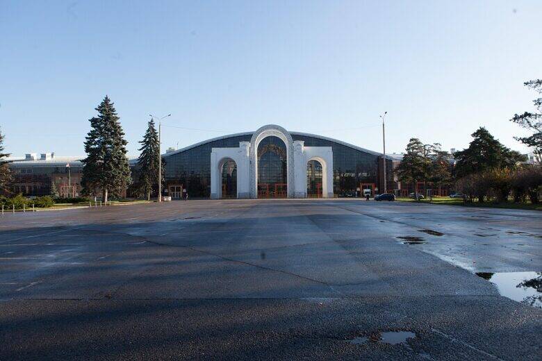 The white North Entrance arch at VDNKh, with its three openings, in front of a glass-fronted pavilion, with a broad square in the foreground.