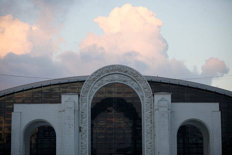The white North Entrance arch at VDNKh, with relief decoration around its central opening, against the backdrop of a glass-fronted pavilion.