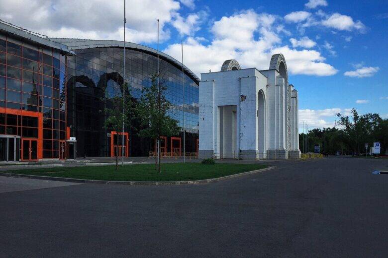 The white North Entrance arch at VDNKh to the right of a glass-fronted pavilion.