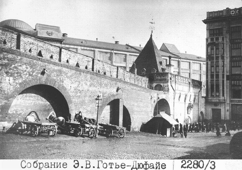 A brick wall with arches; horse-drawn carts and a tent stand in front of it, with people walking in the distance.