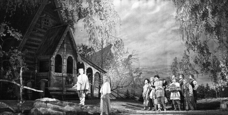 Чёрно-белая театральная сцена: у деревянной постройки стоит одна фигура, справа собралась группа людей в костюмах.
