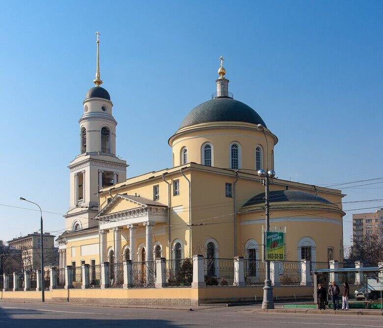 Жёлтый храм с тёмным куполом, белой колокольней и колонным портиком за оградой.