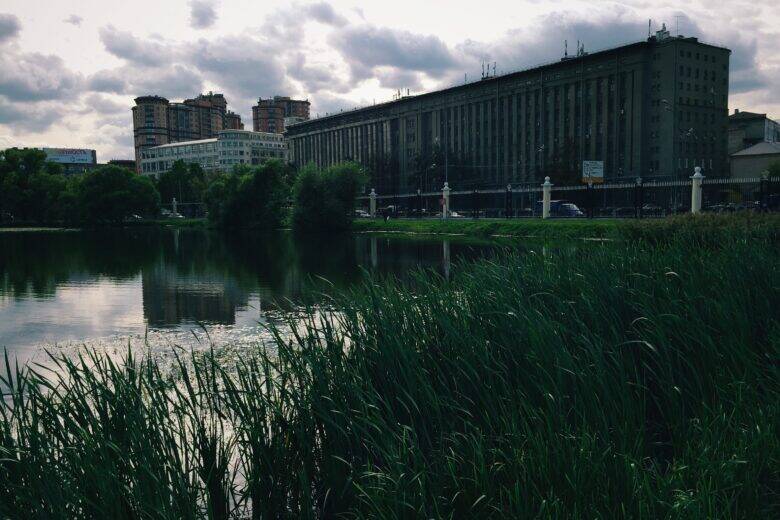 Dense vegetation along the bank and the still surface of a pond; a long building with a tall corner section stands on the far bank.