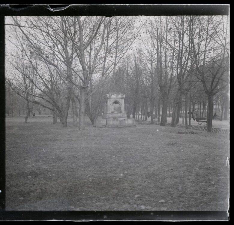 Black-and-white view of the garden: a small stone structure with an arched recess among bare trees; a path and benches to the right.