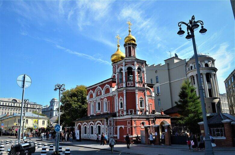 Красно-белая церковь с золотыми главами у пешеходного перехода; перед ней идут прохожие.
