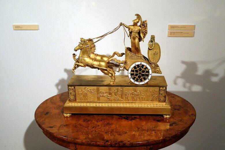 A gilded clock with its dial set in a chariot, a helmeted figure and two horses, on a wooden table.