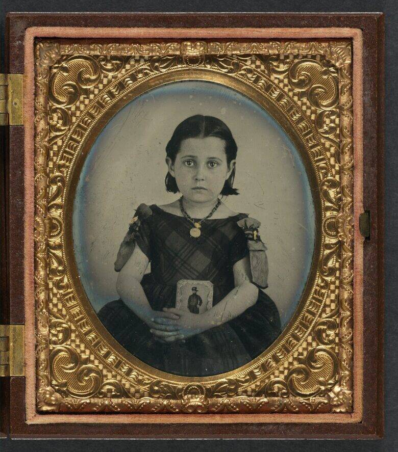 A girl in a dark dress holds a small photographic portrait inside a case decorated with a gold-coloured pattern.