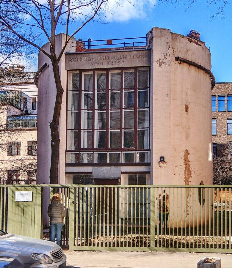 The glazed façade of the Melnikov House between curved walls; a fence and people stand in front of the building.