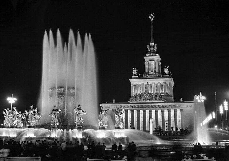 A black-and-white evening view of the Friendship of Peoples fountain: female sculptures around the basin, tall jets of water and the Main Pavilion of the All-Union Agricultural Exhibition behind.