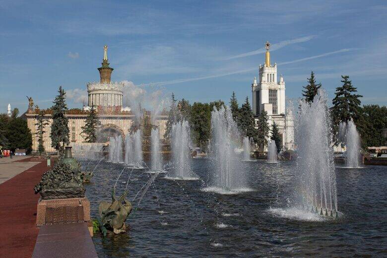 A sculptural group on a pedestal stands at the edge of the pool on the left; behind it are the fountain's water jets and the pavilions of VDNKh.