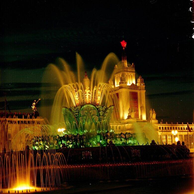 A night-time view of the Stone Flower fountain: the flower in the centre, arcs of water jets, the pool and an illuminated pavilion behind.
