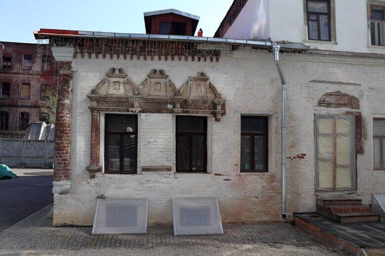 The whitewashed façade of a historic house with two windows in ornate surrounds and patches of exposed red brickwork