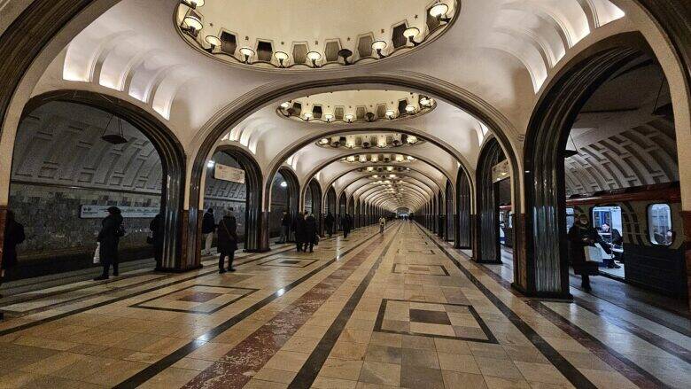 Перспектива зала станции «Маяковская» с арками, потолочными светильниками, пассажирами и поездом у платформы.