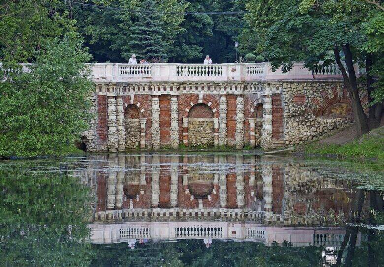 A brick and stone grotto with arches and a white balustrade is reflected in a pond in Lefortovo Park.