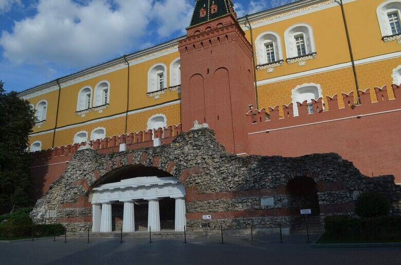 The grotto’s white columned portico beneath a stone arch; the red Kremlin wall and a tower behind it.