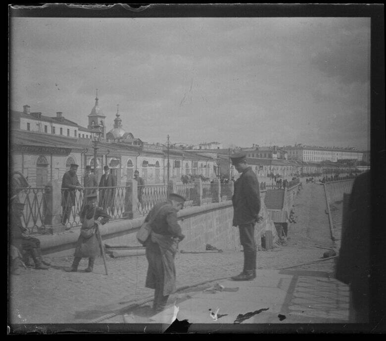 In an old photograph, a man bends down beside a stone parapet in front of a standing person; pedestrians walk along the embankment.