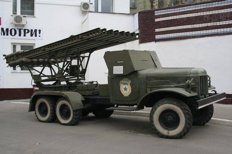 A Katyusha rocket launcher mounted on a truck in a courtyard beside pale buildings.