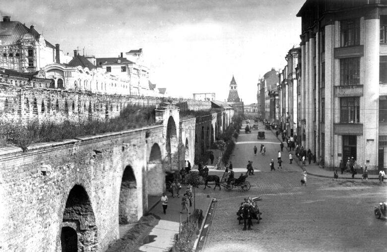 A view along the Kitay-gorod Wall with its arched openings; pedestrians and horse-drawn carriages move along the street, with a tower visible in the distance.