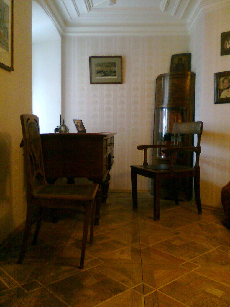 A bright museum room with a wooden table, two chairs and paintings on the walls.
