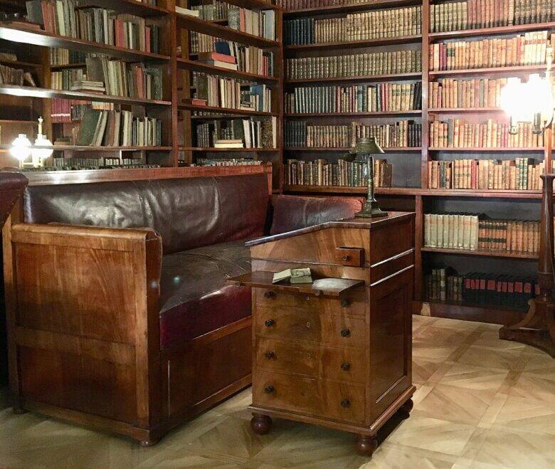 A wooden standing desk with drawers stands beside a dark leather sofa, with bookshelves in the background.