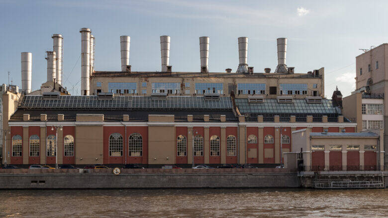 A red-and-beige power station building with arched windows, chimneys behind it and a river in the foreground