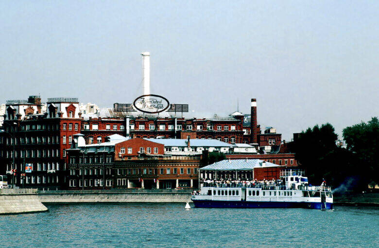 The red-brick buildings of Krasny Oktyabr across the Moskva River; a riverboat passes in front of the embankment.