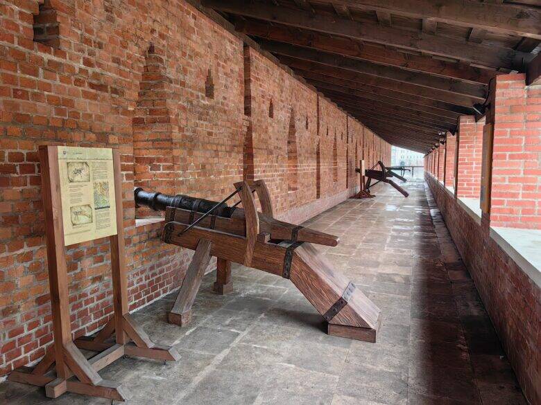 A covered passage along a long brick wall; wooden exhibits and an information board stand on the stone floor.