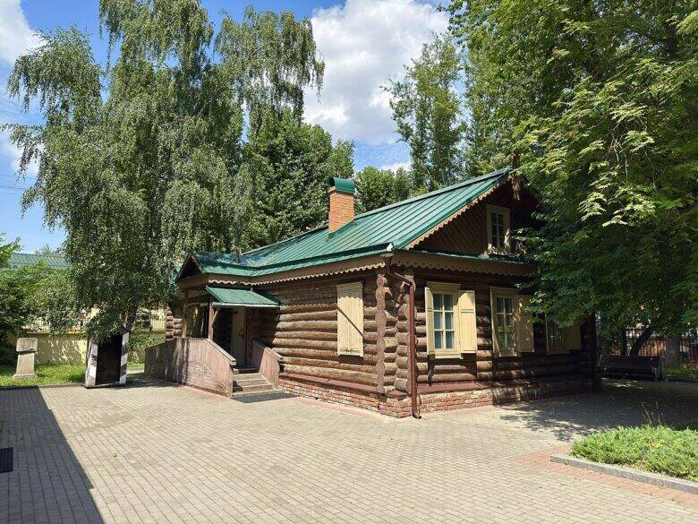Бревенчатая Кутузовская изба с зелёной крышей и светлыми ставнями среди деревьев.
