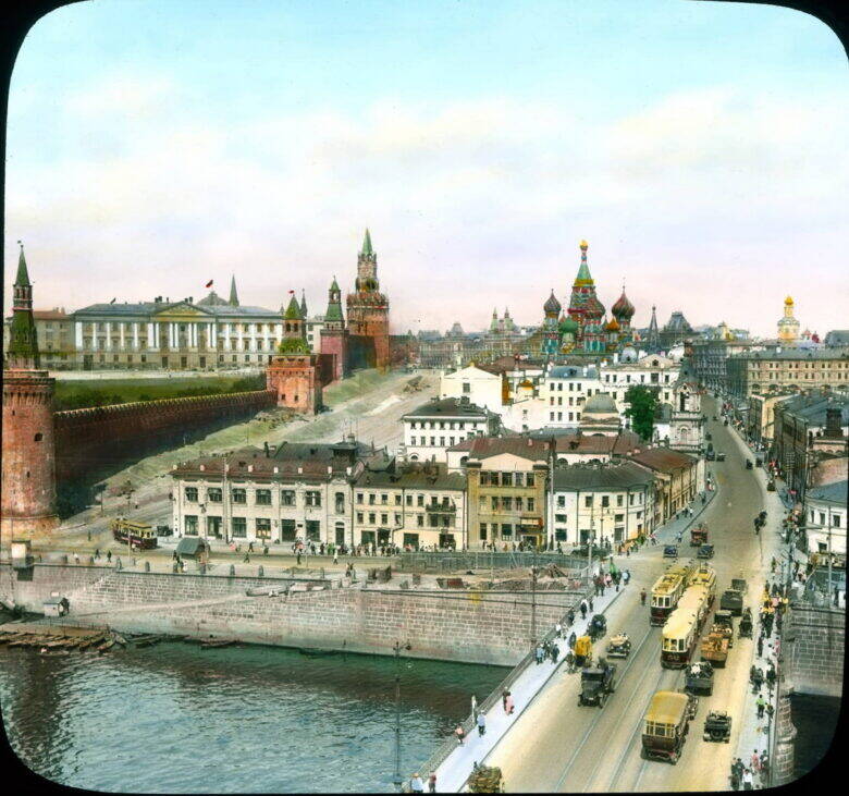 A colourised historical view of Moscow: a bridge with trams, a block of buildings beside the river, the Kremlin wall and the Cathedral of the Intercession.