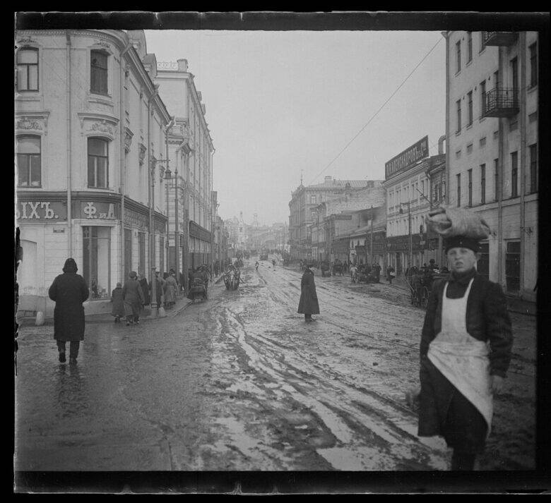 A black-and-white photograph of a street lined with buildings, with passers-by and carts on the wet paving.