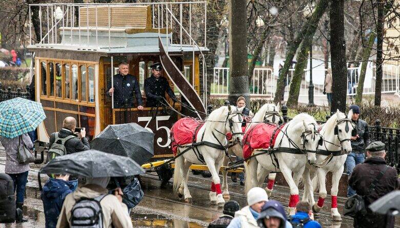 White horses pull a wooden double-decker tram along a wet street; spectators stand around it holding umbrellas.