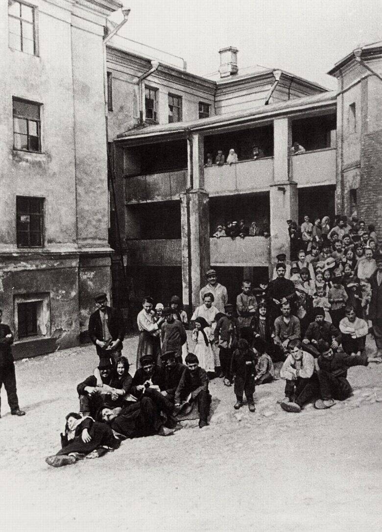 People sitting and standing in a courtyard in front of a building’s open galleries in a historical photograph.