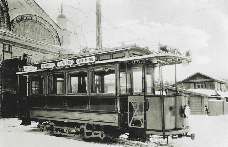 Electric tram No. 19 beside the depot building with its arched entrance; snow covers the ground, and overhead wires run above the tram.