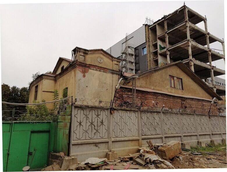 A yellowed historic house behind a fence; plaster has crumbled from the façades, and construction rubble lies in the foreground.