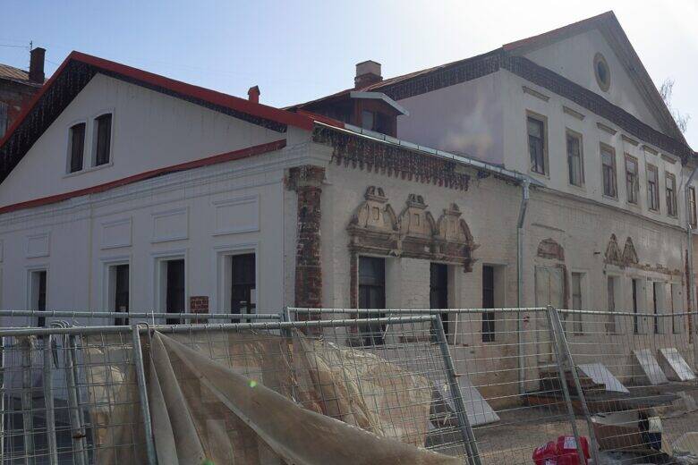 The two-storey façade of a historic house with a round window in the pediment and an adjoining low wing behind construction fencing.