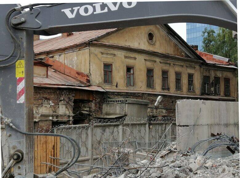 The yellow façade of a historic house behind fencing; an excavator boom and construction rubble are in the foreground.