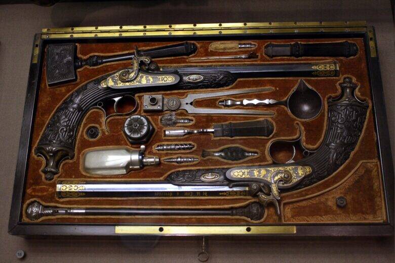 Two ornate pistols and their accessories are arranged in the compartments of an open case with a brown lining.