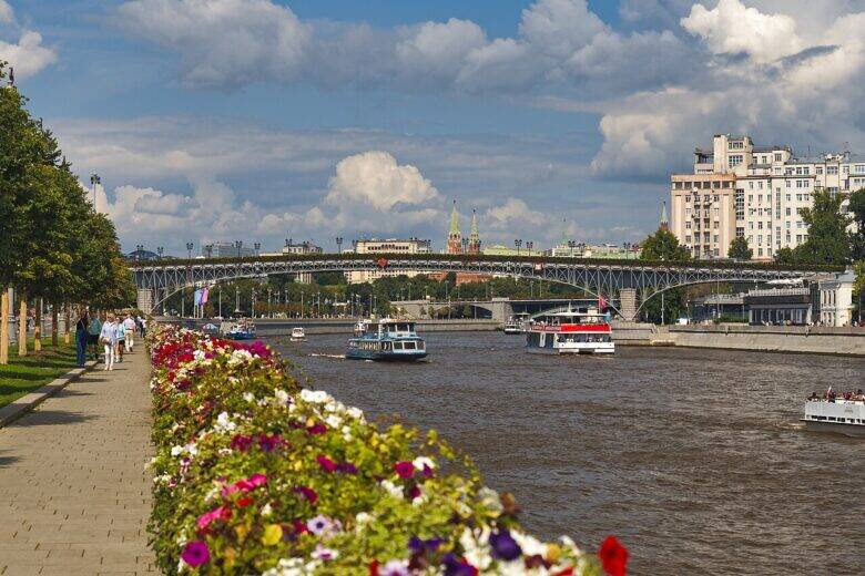 A latticework footbridge crosses the Moskva River; flowerbeds bloom along the bank, and boats pass on the water.