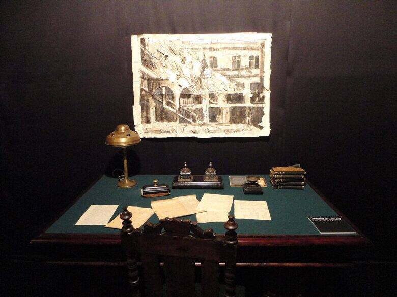A writing desk with papers, books and a lamp against a dark wall in a museum display.