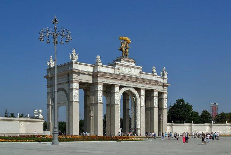 The Main Entrance arch at VDNKh, topped by a sculptural group holding a sheaf, with a lamp post on the left and visitors in front of the arch.
