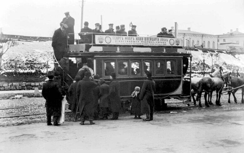People at the entrance to a double-decker horse-drawn tram; passengers are seated on the upper deck, with harnessed horses standing to the right.