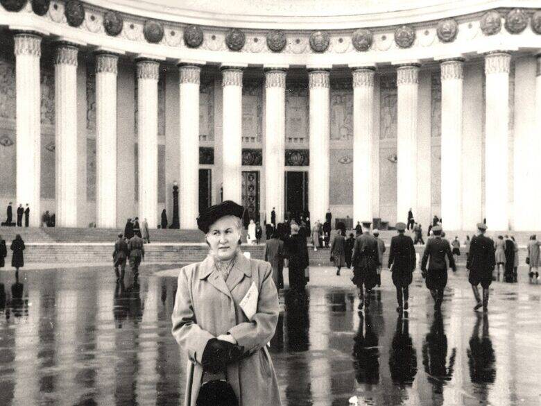 A woman with a bag stands on a wet square in front of a curved colonnade; exhibition visitors walk in the background.
