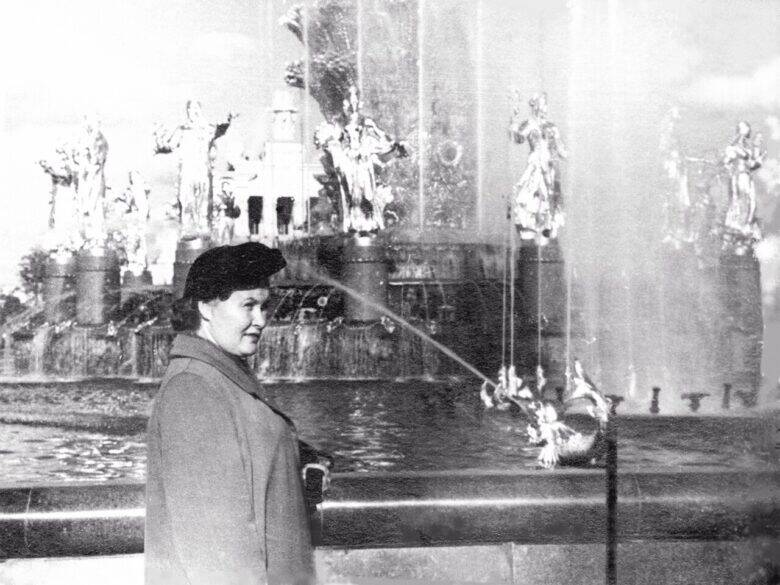 A woman in a coat stands at the edge of the fountain; behind her are female sculptures and tall jets of water.