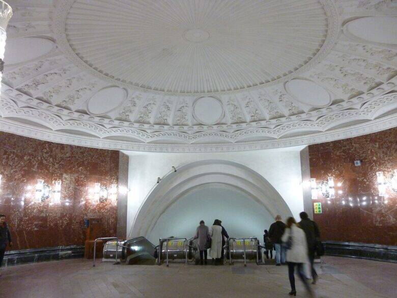 A circular hall with a white dome decorated with plasterwork and red walls; passengers descend towards the escalators.